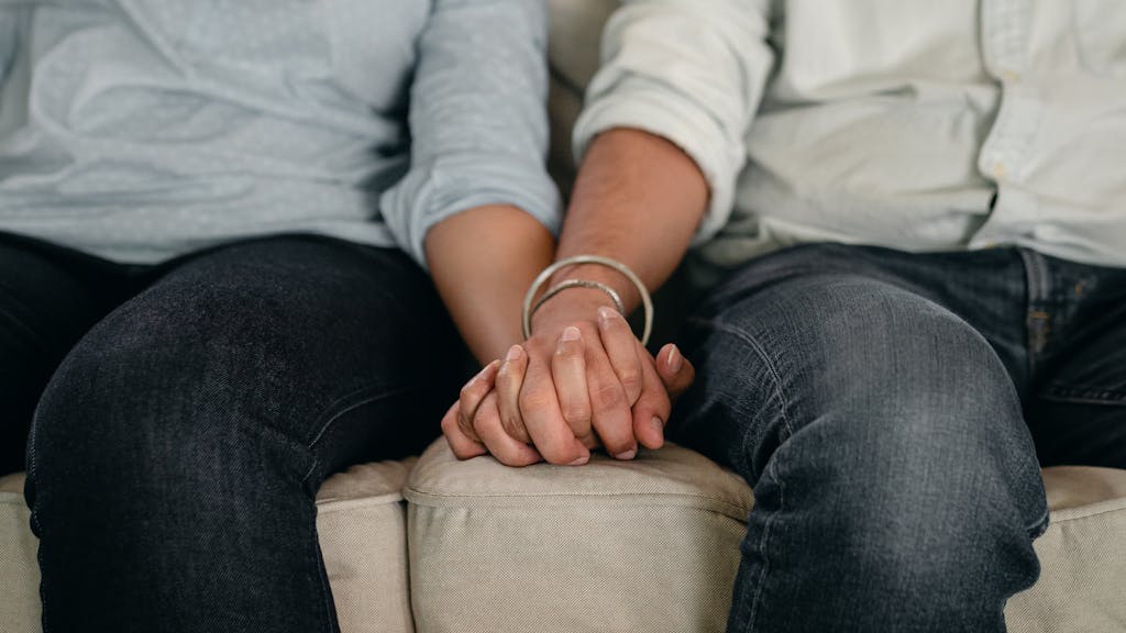 Crop anonymous man in jeans and shirt with wristband holding females hand while resting together on cozy couch in apartment